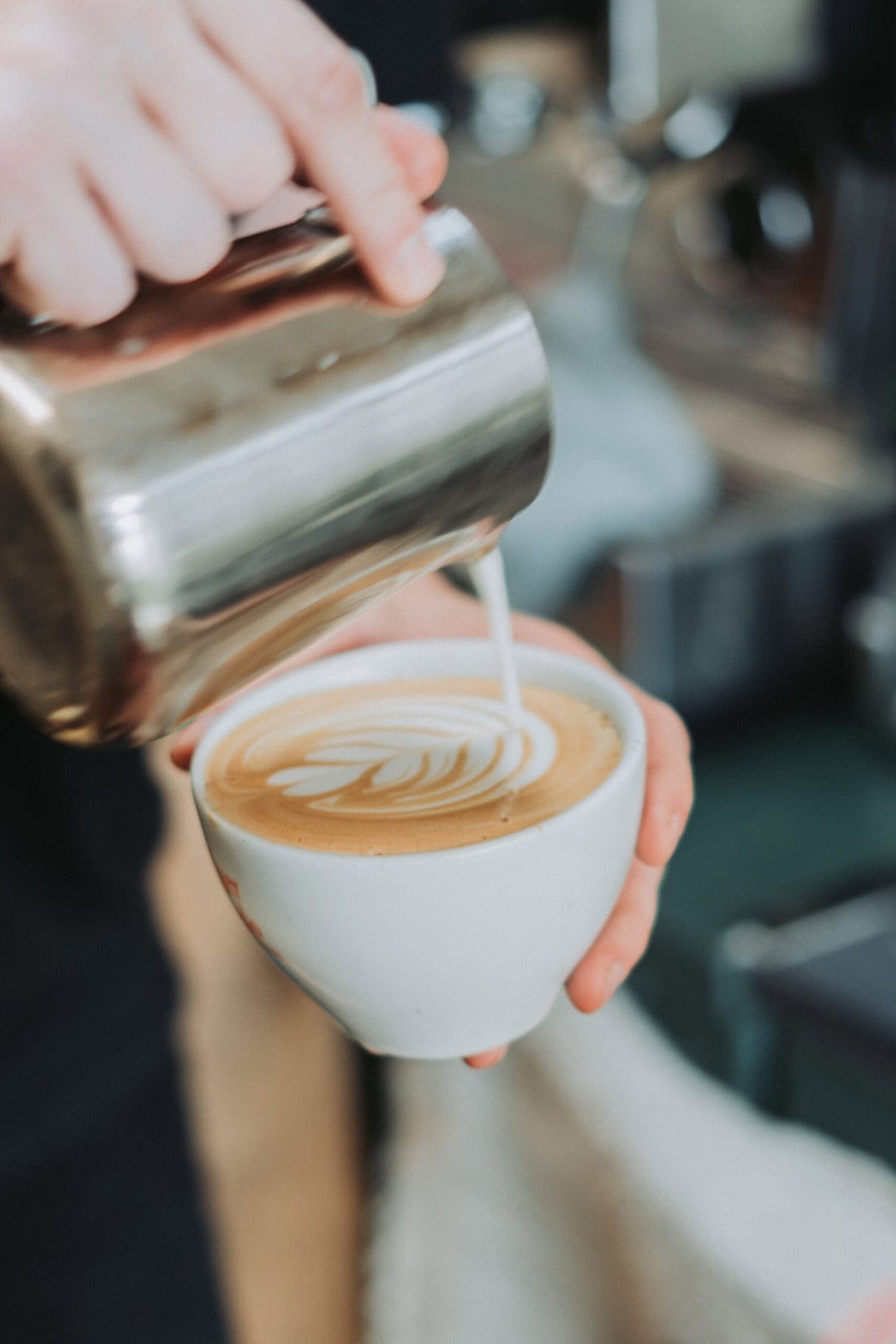 Treehouse coffee. A person is pouring milk into a cup of coffee, creating latte art with a leaf-like pattern on the surface. The coffee cup is white and the background is blurred.