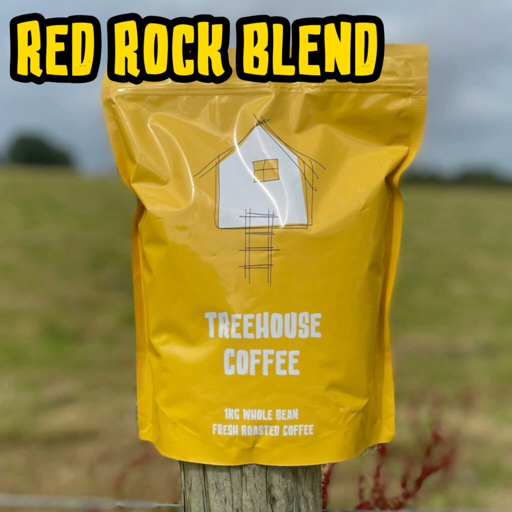 Treehouse coffee. A yellow bag of Treehouse Coffees Red Rock Blend is perched on a wooden post in front of a blurred grassy field. The bag features a simple house illustration and text indicating 1kg of fresh roasted, whole bean coffee.