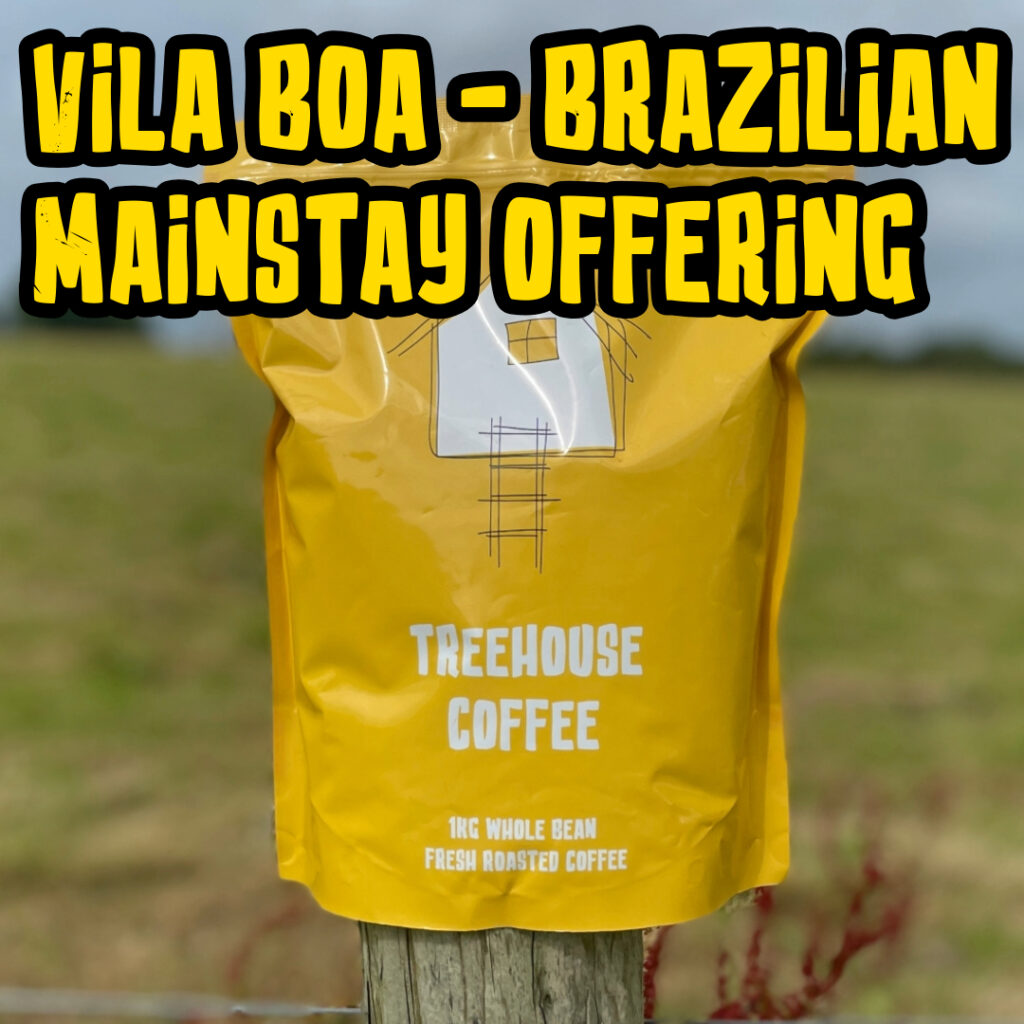 Treehouse coffee. A yellow bag of Treehouse Coffees Vila Boa Brazilian MainstayOffering is perched on a wooden post in front of a blurred grassy field. The bag features a simple house illustration and text indicating 1kg of fresh roasted, whole bean coffee.