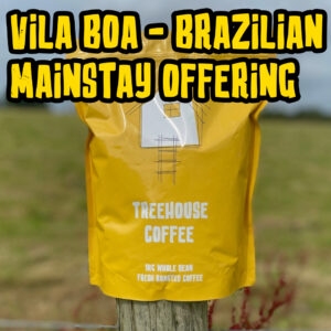 Treehouse coffee. A yellow bag of Treehouse Coffees Vila Boa Brazilian MainstayOffering is perched on a wooden post in front of a blurred grassy field. The bag features a simple house illustration and text indicating 1kg of fresh roasted, whole bean coffee.