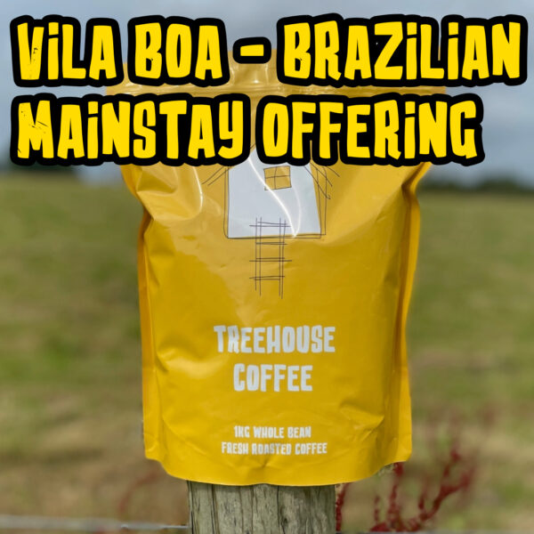 Treehouse coffee. A yellow bag of Treehouse Coffees Vila Boa Brazilian MainstayOffering is perched on a wooden post in front of a blurred grassy field. The bag features a simple house illustration and text indicating 1kg of fresh roasted, whole bean coffee.