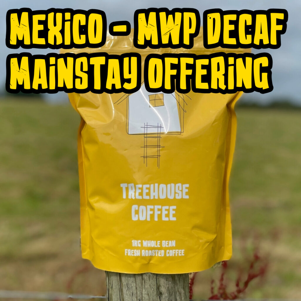 Treehouse coffee. A yellow bag of Treehouse Coffees Mexico MWP Decaf Mainstay Offering is perched on a wooden post in front of a blurred grassy field. The bag features a simple house illustration and text indicating 1kg of fresh roasted, whole bean coffee.