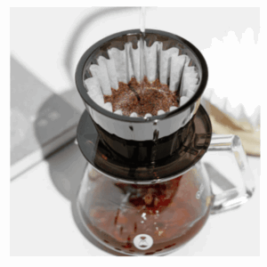 A top-down view of a pour-over coffee setup with hot water being poured over ground coffee in a paper filter inside a black plastic dripper, which is placed on top of a glass carafe partially filled with brewed coffee.
