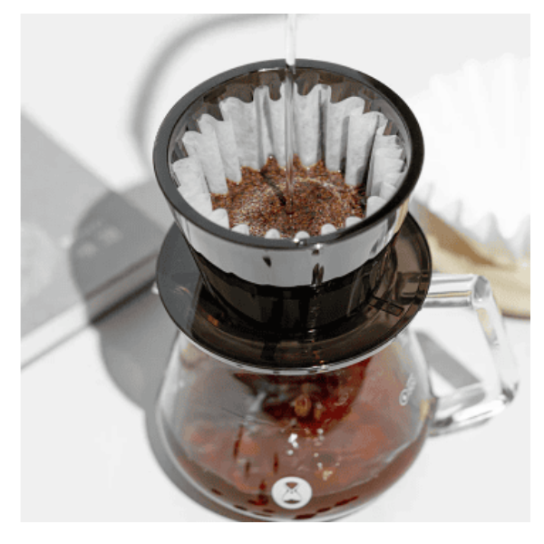 A top-down view of a pour-over coffee setup with hot water being poured over ground coffee in a paper filter inside a black plastic dripper, which is placed on top of a glass carafe partially filled with brewed coffee.