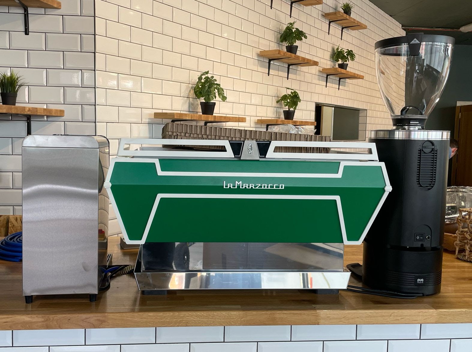 A green La Marzocco espresso machine on a wooden counter in a modern café. To the left is a stainless steel water filtration unit, and to the right is a black coffee grinder. The background features white subway tile walls with small potted plants on wooden shelves.