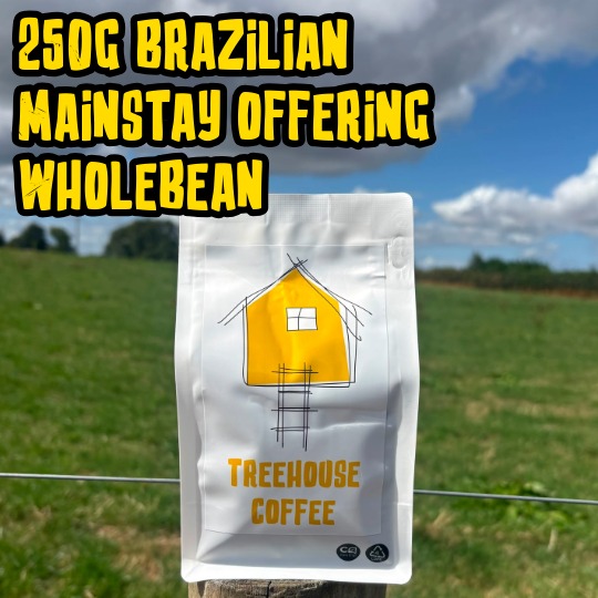Treehouse coffee. A yellow bag of Treehouse Coffees Vila Boa Brazilian MainstayOffering is perched on a wooden post in front of a blurred grassy field. The bag features a simple house illustration and text indicating 250g of fresh roasted, whole bean coffee.